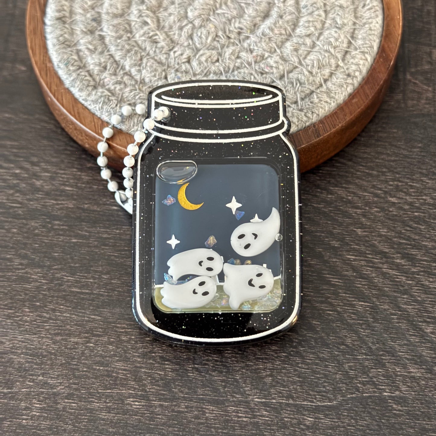 A mason jar-shaped pendant with a black glittery background, white ghosts, a yellow crescent moon, and small star details, hanging from a white beaded chain.