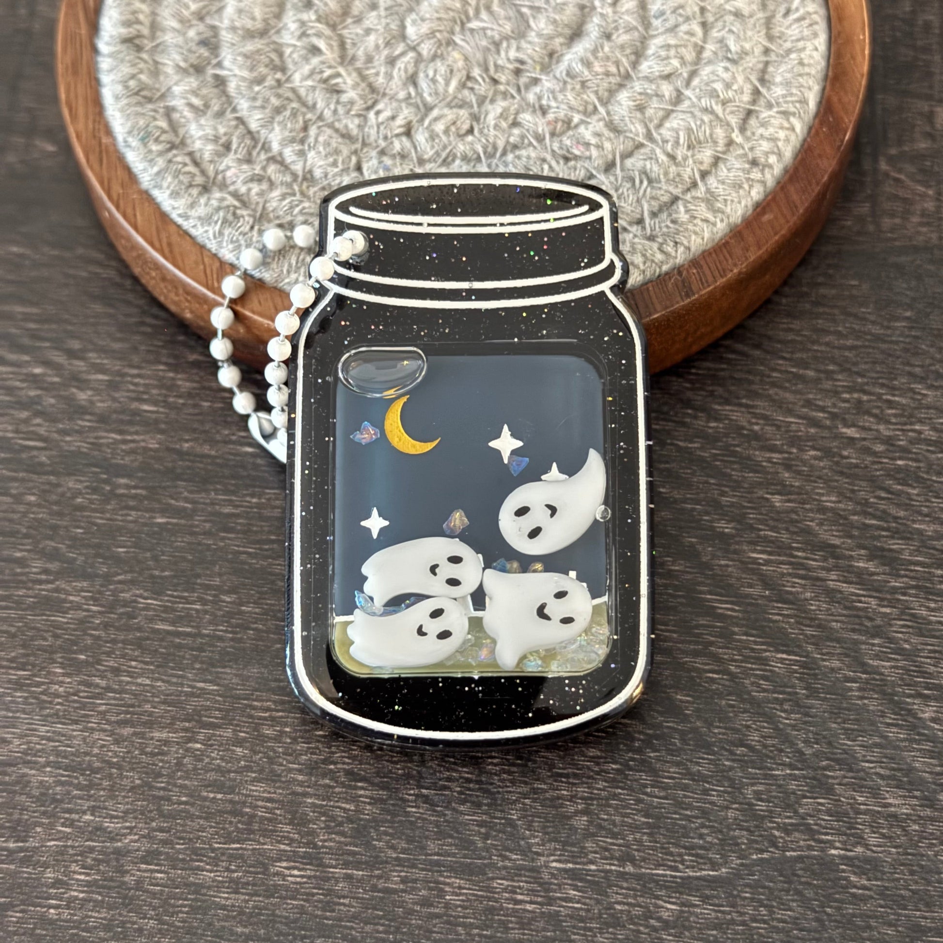 A mason jar-shaped pendant with a black glittery background, white ghosts, a yellow crescent moon, and small star details, hanging from a white beaded chain.