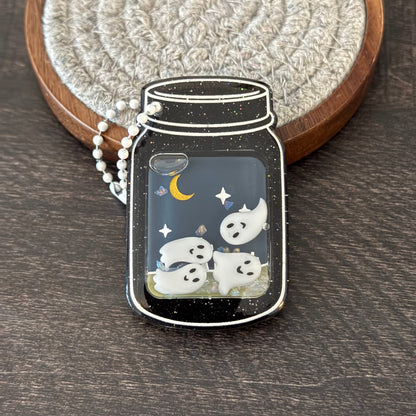 A mason jar-shaped pendant with a black glittery background, white ghosts, a yellow crescent moon, and small star details, hanging from a white beaded chain.